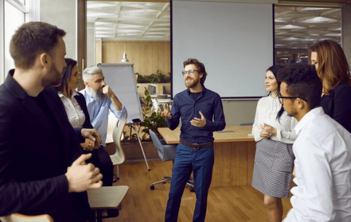 Une équipe de professionnels en pleine discussion collaborative dans un bureau moderne. La salle est lumineuse grâce à de grandes fenêtres et est équipée de mobilier contemporain. Un tableau blanc sur pied est visible, suggérant une session interactive de présentation ou de brainstorming. Les visages des participants sont floutés pour des raisons de confidentialité. Cette image est idéale pour illustrer des thèmes liés aux réunions d’affaires, à la collaboration en équipe, aux environnements d’entreprise ou aux interactions en milieu de travail